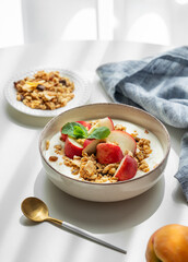 Natural greek yogurt with granola or muesli and peach in a bowl on a white table near window
