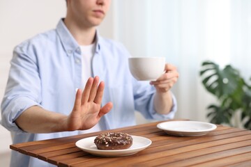 Gluten free diet. Man with cup of drink refusing from donut at wooden table indoors, closeup
