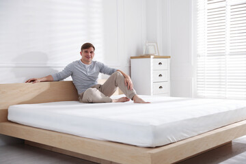 Young man sitting on bed with soft mattress at home, space for text