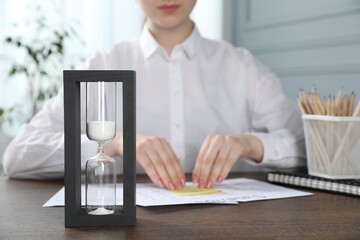 Deadline. Woman attaching sticky note to calendar at wooden table in office, focus on hourglass