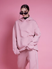 A woman in a pink hoodie and sweatpants poses for a photo. The pink color of her outfit gives off a...