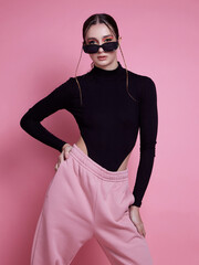 A woman in a pink hoodie and sweatpants poses for a photo. The pink color of her outfit gives off a...