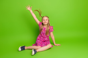Preteen girl in a bright floral dress sitting on a vibrant background, smiling joyfully with an arm raised in excitement.