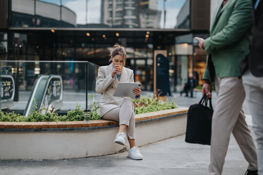 Businesswoman multitasking, seated outdoors in a modern cityscaped plaza, drinking coffee, utilizing a tablet for work, amidst a dynamic backdrop featuring people and office buildings during the day.