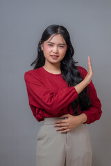 Young Indonesian woman wearing a red top and beige pants, raising her hand in a “stop” gesture...