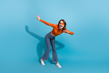 Young woman with orange top posing in playful gesture against vibrant blue background, highlighting...