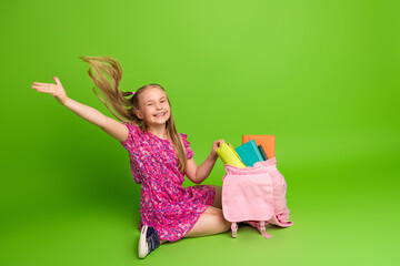 Cheerful young schoolgirl in a pink dress opening her backpack, surrounded by book, with a vibrant...