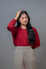An Indonesian woman with a red top and beige pants is touching her head, showing a facial...