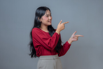 An Indonesian Asian woman wearing a red long-sleeve shirt and beige pants, smiling and pointing...