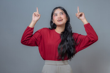 Young Indonesian woman in a red shirt and beige pants, smiling while pointing upwards with both...
