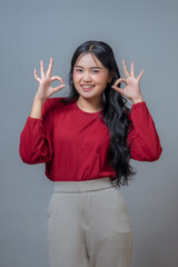 Charming young Indonesian woman in a red top and beige pants making a double “OK” hand gesture...