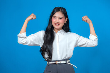 Confident young Asian woman smiling and flexing both arms to show strength and empowerment, wearing a white shirt and gray pants, posing against a clean blue background in studio.