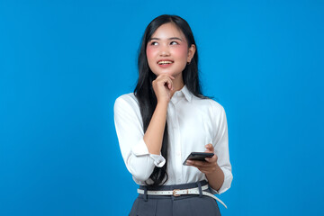 Young Asian woman in white shirt holding smartphone while smiling and looking up with thoughtful...
