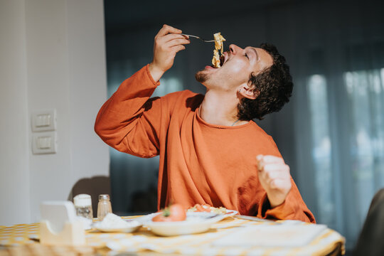 A young man appears to relish a delicious pasta meal while sitting at a table in the cozy ambiance of a home kitchen. - Powered by Adobe