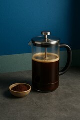 A clear glass French press filled with rich, dark coffee next to a bowl of coffee ground