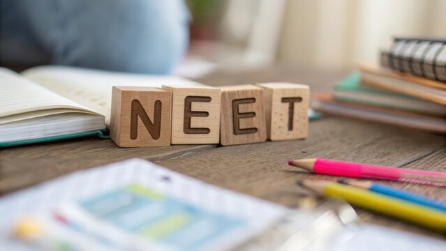Soft focus scene of wooden letter blocks arranged to form the word NEET with study materials

