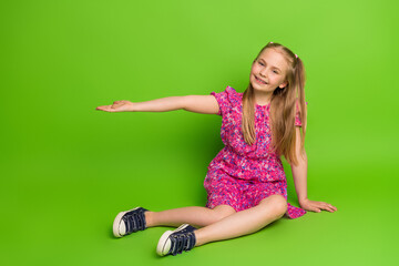 Cute girl smiling while gesturing with hand on vibrant green background in a colorful dress and classic sneakers