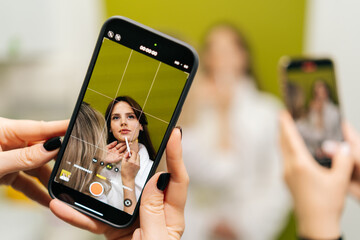 Female hands holding smartphone recording video of beautician applying makeup on model face, creating content for social media or online tutorial in cosmetology clinic. Concept of beauty treatment.