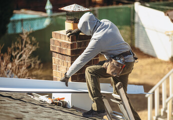 A roofer in a hoodie is working on a roof, installing or repairing a section near a chimney with...