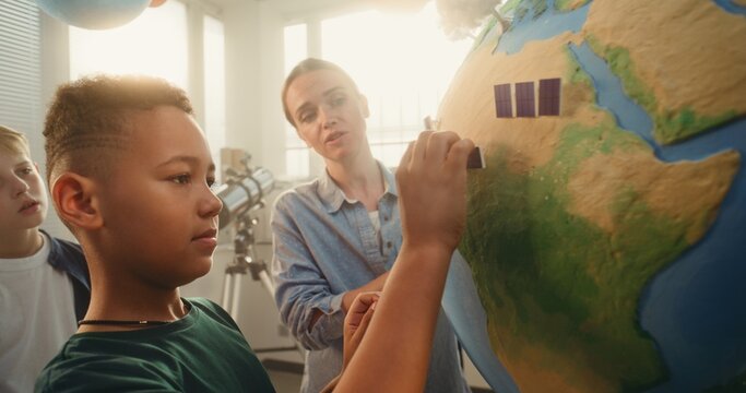 Science STEM Lesson: Female Teacher Educating Renewable Energy to Team of Smart Kids in Multiethnic Classroom. African American School Boy Attaching Miniature Solar Panel to Earth Model. Close Up.