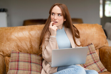 Young woman sitting on a sofa, looking thoughtfully at a laptop while pondering an important...