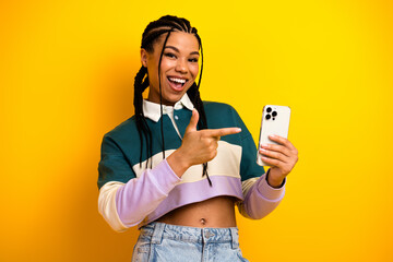 Cheerful young woman with braided hair holding a smartphone against a vibrant yellow background...