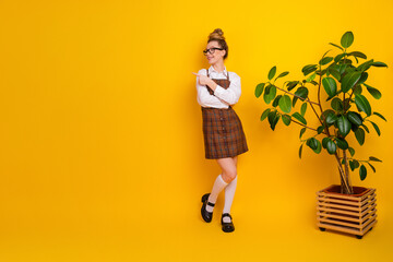 Cute woman in stylish checkered uniform stands indoors pointing sideways with a rubber plant in a...
