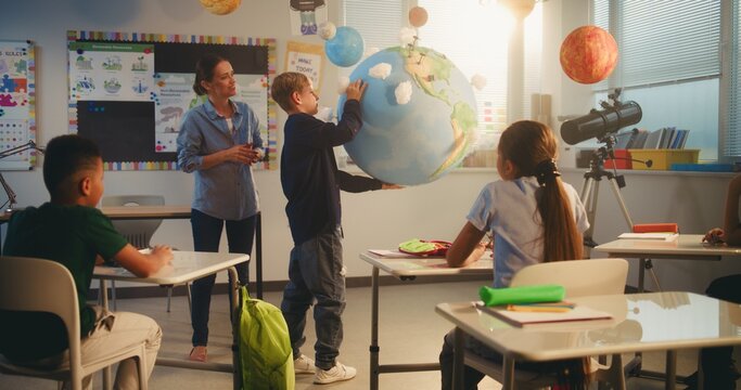 Primary School Boy Presenting Green Energy Sources in Front of Class Using Earth Model. Female Teacher Educating Smart Kids During Science Lesson in Multiethnic Classroom. STEM Education. Dolly Shot. - Powered by Adobe