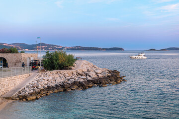 Small marina with rocky coast and yacht floating near a walkway with bench. Calm sea under early...