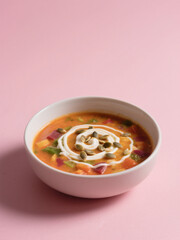 A vibrant bowl of creamy vegetable soup with a swirl of cream and pumpkin seeds, served on a pastel pink background.