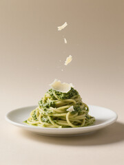 A plate of spaghetti with pesto sauce, topped with Parmesan cheese shavings falling mid-air, set against a neutral beige background.