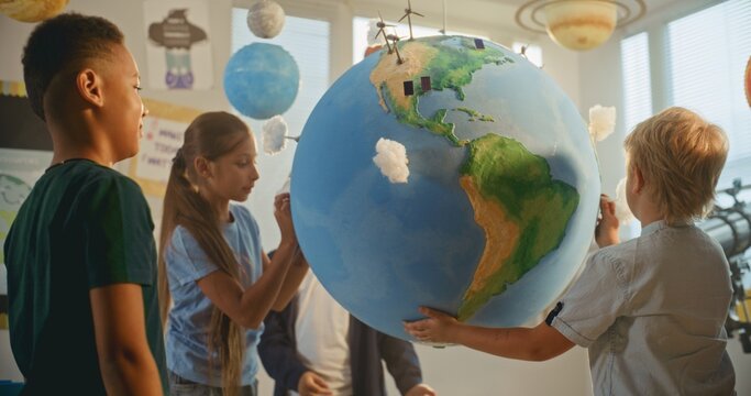 Renewable Energy STEM Class: Elementary School Students Attaching Miniature Models of Clouds to Globe. Female Teacher Explaining Science and Energy Sources to Smart Diverse Kids in Modern Classroom.