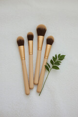 Three natural makeup brushes with wooden handles and soft bristles neatly arranged on a white towel, complemented by a small green leaf for a fresh, minimalistic composition.