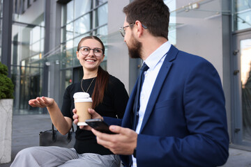 Happy coworkers with smartphone and coffee discussing something near office building