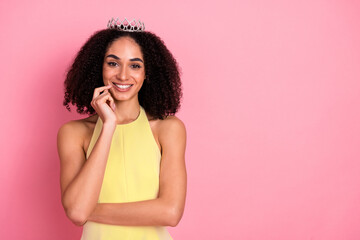 Smiling young woman wearing a yellow dress and crown standing against a pink background showcasing...