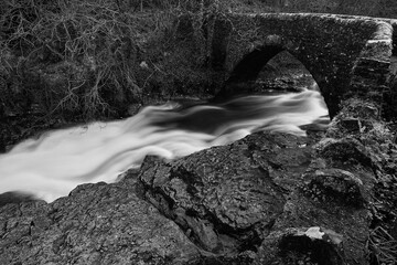 Long exposure view shows the smooth, flowing water of river passing beneath an old stone bridge in...