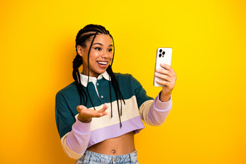 Young woman with braided hair holding a smartphone gestures enthusiastically against a vivid yellow...