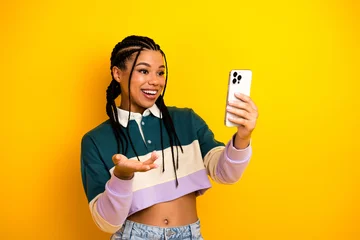  Young woman with braided hair holding a smartphone gestures enthusiastically against a vivid yellow background © deagreez