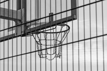 Outdoor metal basketball hoop with mesh net against sunset sky. View from behind fence. Black and white shot