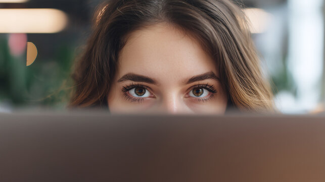 Young woman with tired eyes peeking over laptop screen, showing focus and slight fatigue in blurred indoor setting with soft lighting and natural background