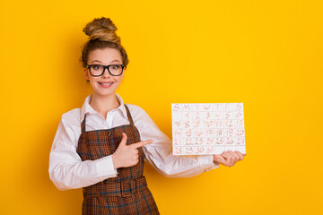 Cheerful young schoolgirl holding and pointing to calendar against yellow background, promoting education and planning