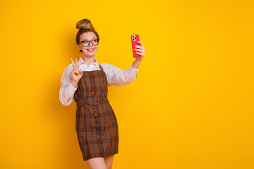 Young student in stylish plaid dress capturing moments on smartphone against vibrant yellow...