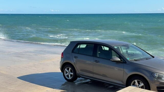 The car is parked dangerously close to the stormy sea.