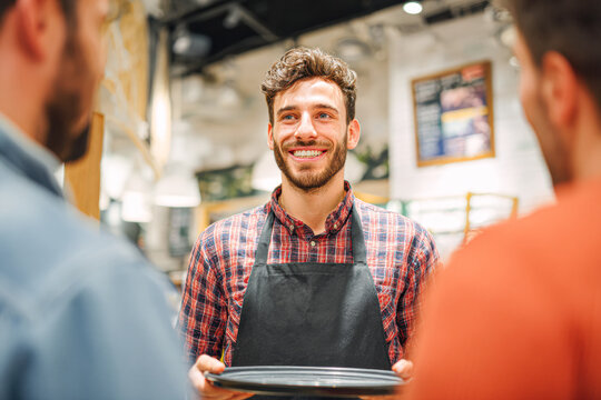 A smiling waiter in a plaid shirt and black apron holds an empty plate, engaging with customers in a cozy, well-lit restaurant setting.