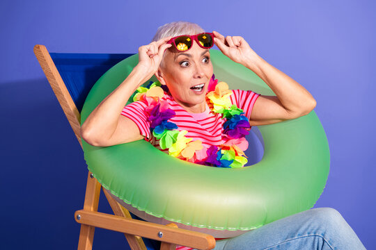 Cheerful senior woman enjoying summer vibes with a green inflatable ring, colorful lei, and striped t-shirt against a vibrant background