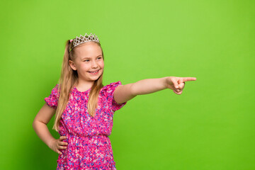 Adorable little girl wearing a floral dress and tiara pointing with confidence on a vibrant green background