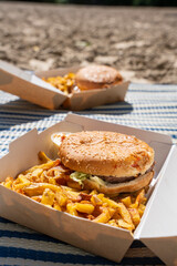 Zoom sur une serviette posée sur le sable avec des burgers dans leur emballage carton accompagnés de frites, évoquant un pique-nique estival en bord de mer.