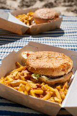 Zoom sur une serviette posée sur le sable avec des burgers dans leur emballage carton accompagnés de frites, évoquant un pique-nique estival en bord de mer.