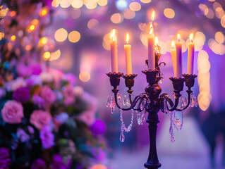 A close-up of a candelabra with lit white candles next to a bouquet of pink and white flowers, all set against a blurry purple background with bokeh lights.