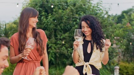 Graceful brunette female in colored dress dancing with curly-haired woman. Holding wine glass. Resting in garden. Laughing during outdoor party surrounded by friends. Recreation concept.